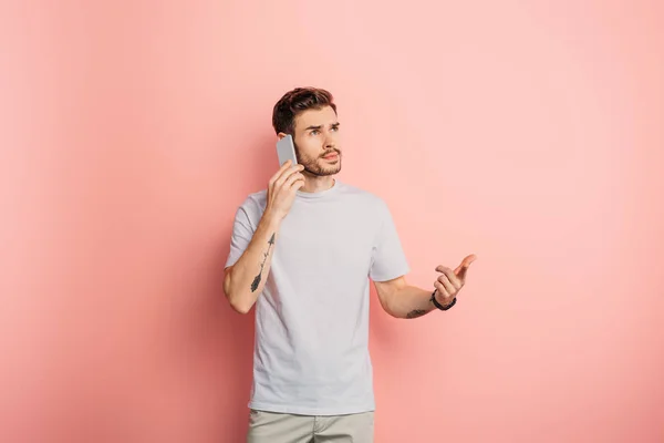 Nachdenklicher junger Mann schaut weg, während er auf rosa Hintergrund mit dem Smartphone spricht — Stockfoto