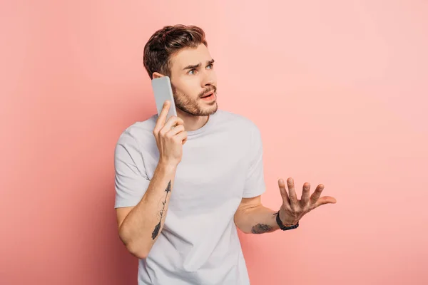 Schockierter junger Mann steht mit offenem Arm da, während er auf rosa Hintergrund mit dem Smartphone spricht — Stockfoto
