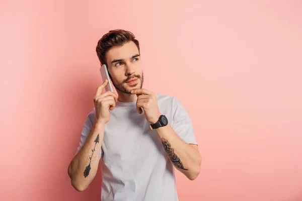 Pensive young man touching chin while talking on smartphone on pink background — Stock Photo