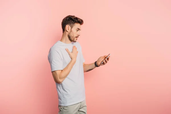 Удивленный молодой человек трогает грудь, держа смартфон на розовом фоне — стоковое фото