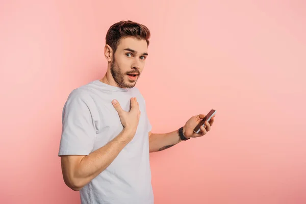 Scioccato giovane uomo toccando petto tenendo smartphone su sfondo rosa — Foto stock