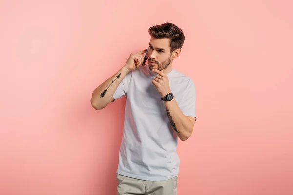 Thoughtful young man touching chin while talking on smartphone on pink background — Stock Photo