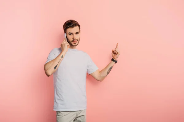 Ernster junger Mann zeigt Ideengeste, während er auf einem Smartphone auf rosa Hintergrund spricht — Stockfoto