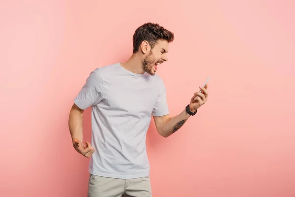 Irritated young man screaming while having video call on smartphone on pink background — Stock Photo