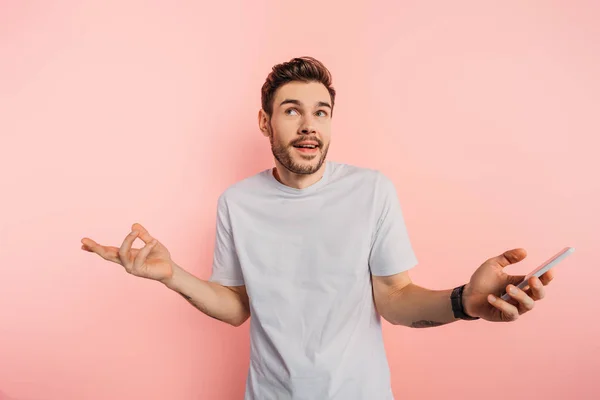 Confused young man showing shrug gesture while holding smartphone on pink background — Stock Photo
