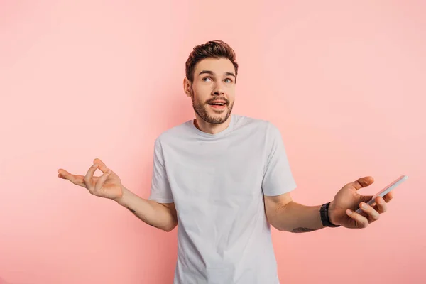 Joven confundido mostrando gesto encogiéndose de hombros mientras sostiene el teléfono inteligente sobre fondo rosa - foto de stock