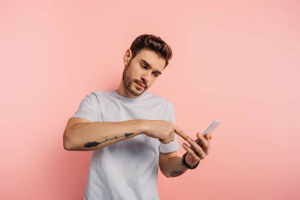 Впевнений молодий чоловік вказує пальцем на смартфон під час відеодзвінка на рожевому фоні — стокове фото
