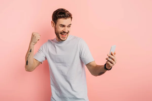 Aufgeregter junger Mann zeigt Siegergeste bei Videoanruf auf Smartphone auf rosa Hintergrund — Stockfoto