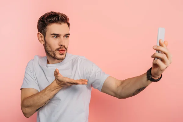 Guapo joven soplando aire beso mientras tiene vídeo chat en el teléfono inteligente sobre fondo rosa — Stock Photo