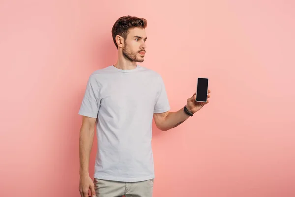 Jeune homme confiant montrant smartphone avec écran vide et regardant loin sur fond rose — Photo de stock
