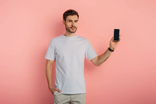 Handsome confident man with hand in pocket showing smartphone with blank screen on pink background — Stock Photo
