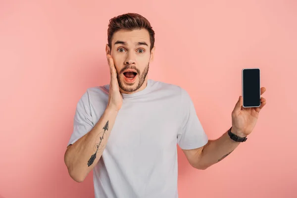 Schockierter Mann berührt Gesicht, während er Smartphone mit leerem Bildschirm auf rosa Hintergrund zeigt — Stockfoto