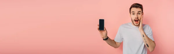 Панорамный снимок шокированного человека, трогательного лица, показывающего смартфон с пустым экраном на розовом фоне — стоковое фото
