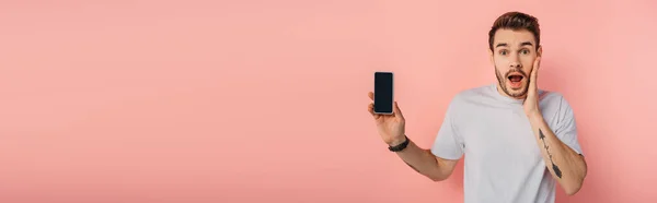 Panoramic shot of shocked man touching face while showing smartphone with blank screen on pink background — Stock Photo