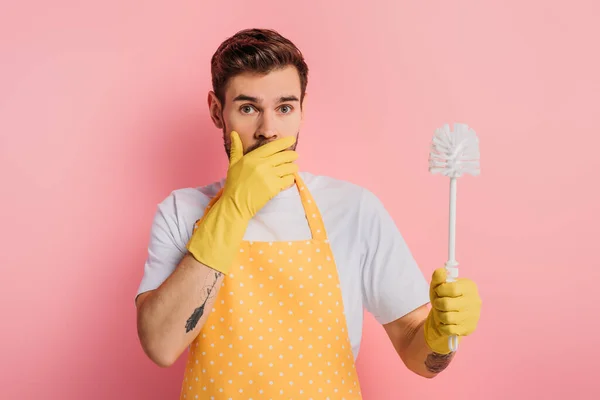 Schockierter junger Mann in Schürze und Gummihandschuhen, der Mund mit der Hand verdeckt, während er Toilettenbürste auf rosa Hintergrund hält — Stockfoto