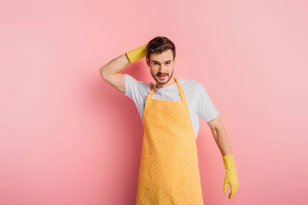 Jeune homme souriant en tablier et gants en caoutchouc touchant la tête tout en regardant la caméra sur fond rose — Photo de stock