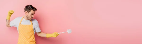 Panoramic shot of young man in apron and rubber gloves imitating fencing with toiltet brush on pink background — Stock Photo