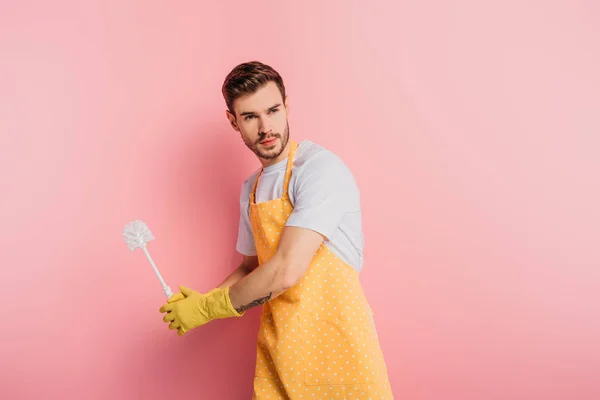 Concentrated young man in apron and rubber gloves imitating playing baseball with toliet brush on pink background — Stock Photo