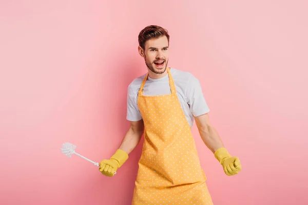 Jeune homme irrité en tablier et gants en caoutchouc criant tout en tenant la brosse de toilette sur fond rose — Photo de stock