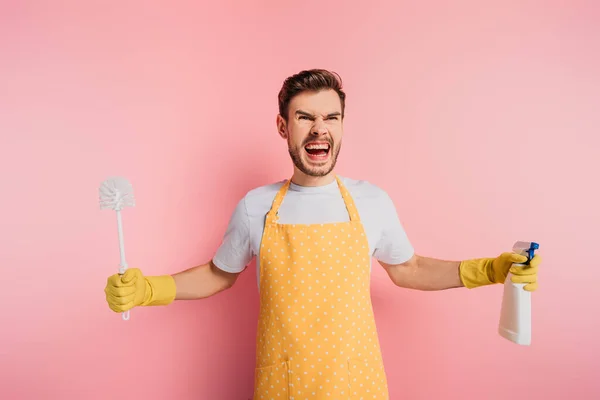 Jeune homme agressif en tablier et gants en caoutchouc tenant brosse de toilette et vaporisateur sur fond rose — Photo de stock