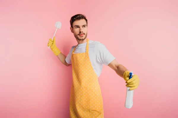 Arrabbiato giovane uomo in grembiule e guanti di gomma guardando lontano mentre tiene bottiglia spray e spazzola da bagno su sfondo rosa — Foto stock