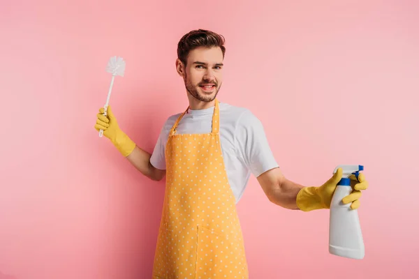 Joyeux jeune homme en tablier et gants en caoutchouc tenant bouteille de pulvérisation et brosse de toilette sur fond rose — Photo de stock