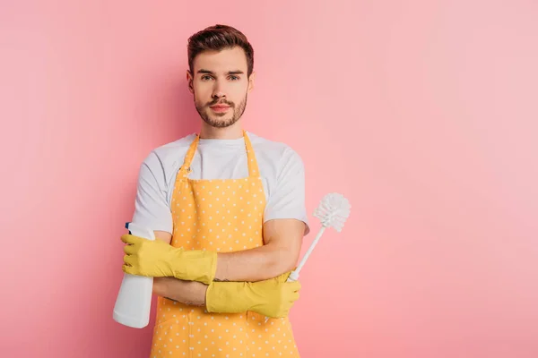 Serious young man in apron and rubber gloves holding spray bottle and toilet brush on pink background — Stock Photo