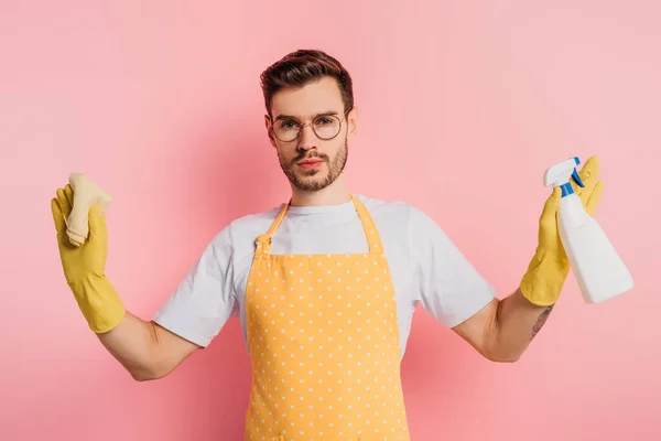 Sérieux jeune homme en tablier et gants en caoutchouc tenant vaporisateur et chiffon sur fond rose — Photo de stock