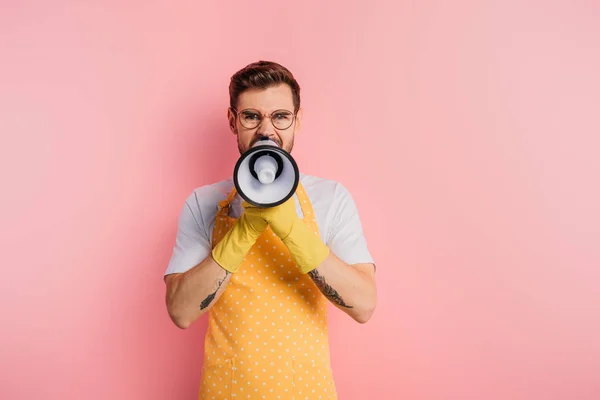 Irritated young man in apron and rubber gloves shouting in megaphone on pink background — Stock Photo