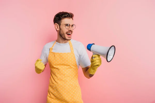 Jeune homme irrité en tablier et gants en caoutchouc criant en mégaphone sur fond rose — Photo de stock