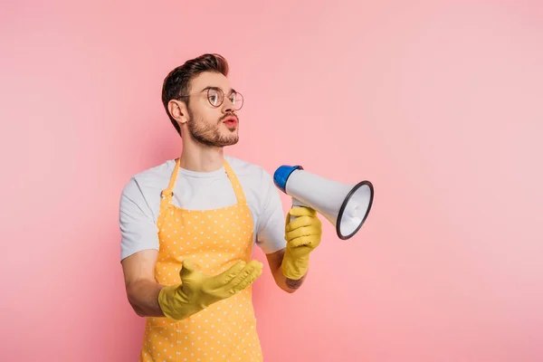 Displeased young man in apron and rubber gloves holding megaphone while standing with open arm on pink background — Stock Photo