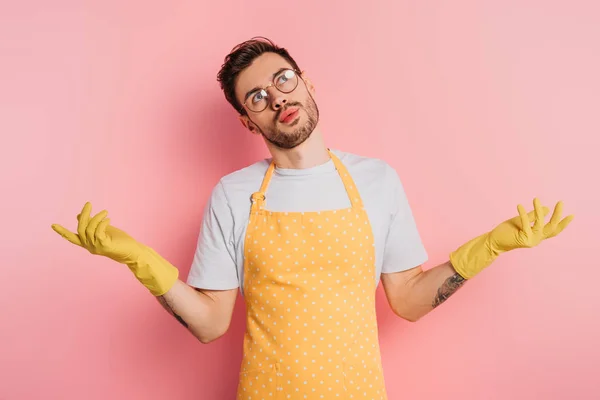 Confuso joven en delantal y guantes de goma mostrando gesto encogiéndose de hombros sobre fondo rosa - foto de stock