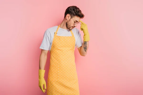 Thoughtful young man in apron and rubber gloves touching forehead with finger on pink background — Stock Photo