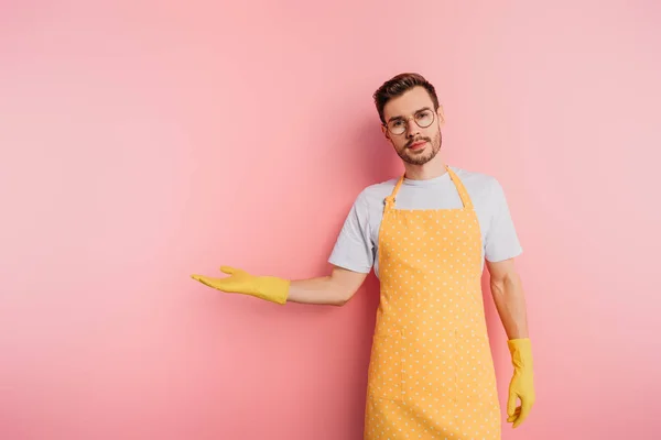 Serious young man in apron and rubber gloves standing with open arm on pink background — Stock Photo
