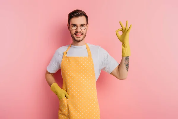 Heureux jeune homme en tablier et gants en caoutchouc montrant geste correct sur fond rose — Photo de stock