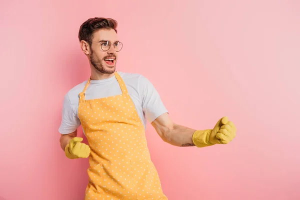 Jeune homme excité en tablier et gants en caoutchouc montrant geste gagnant sur fond rose — Photo de stock