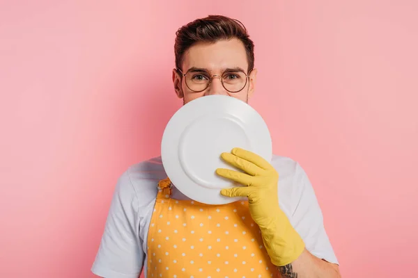 Cheerful young man in apron and rubber gloves covering face with clean plate on pink background — Stock Photo