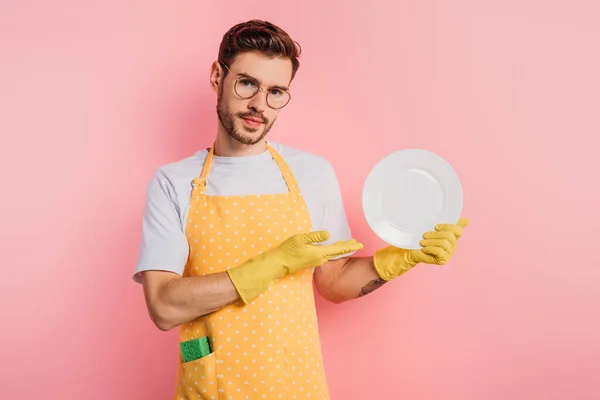 Joven confiado en delantal y guantes de goma que muestran el plato limpio sobre fondo rosa - foto de stock