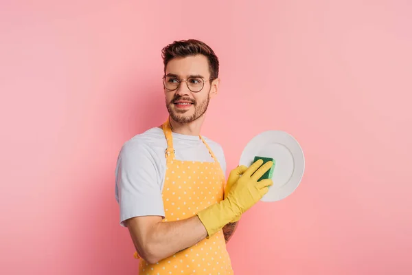 Happy young man in apron and rubber gloves cleaning plate with sponge on pink background — Stock Photo