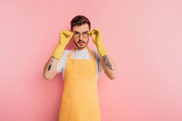 Selbstbewusster junger Mann in Schürze und Gummihandschuhen, der eine Brille auf rosa Hintergrund berührt — Stockfoto
