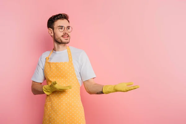 Joven sonriente en delantal y guantes de goma apuntando con las manos sobre fondo rosa - foto de stock