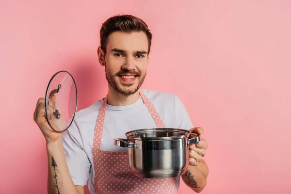Cheerful young man in apron opening saucepan on pink background — Stock Photo