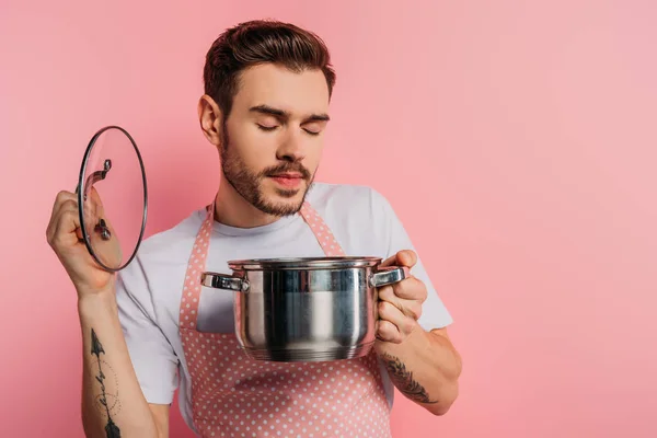 Joven complacido en delantal disfrutando del sabor con los ojos cerrados al abrir la cacerola sobre fondo rosa - foto de stock