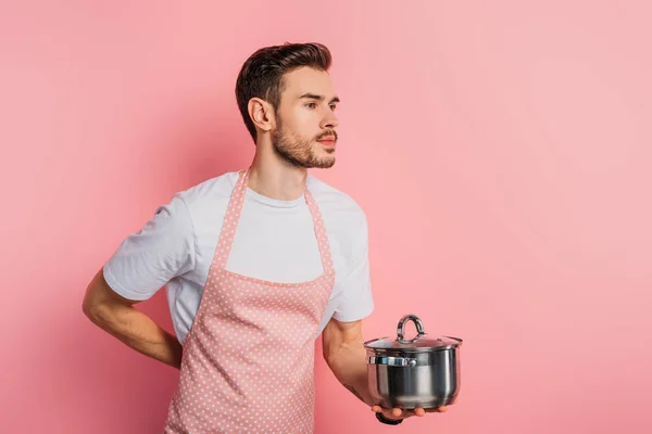 Schöner junger Mann in Schürze imitiert Kellner, während er Kochtopf auf rosa Hintergrund hält — Stockfoto