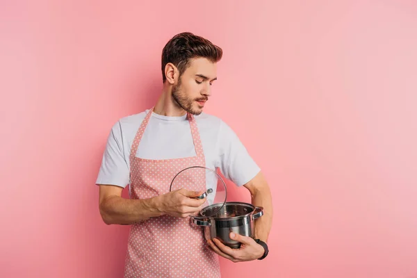 Serious young man in apron opening and look into saucepan on pink background — Stock Photo