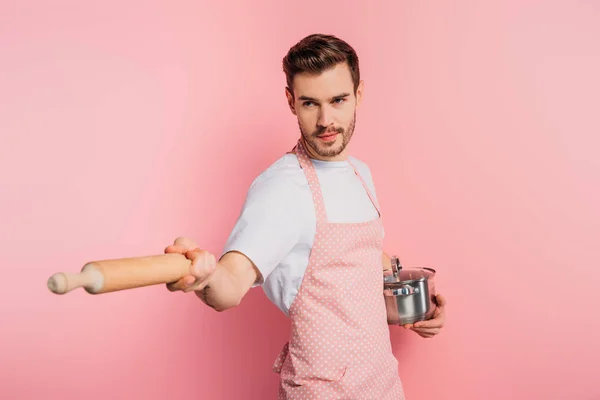 Concentrated young man in apron imitating fencing with saucepan and rolling pin on pink background — Stock Photo