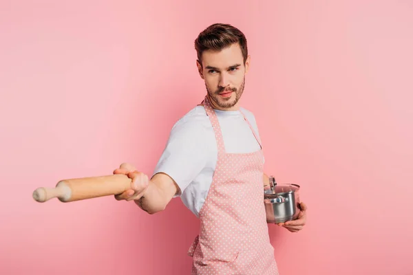 Jeune homme concentré dans tablier imitant l'escrime avec casserole et rouleau à pâtisserie sur fond rose — Photo de stock