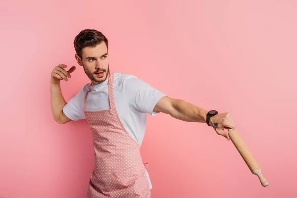 Concentrated young man in apron imitating playing baseball with rolling pin on pink background — Stock Photo