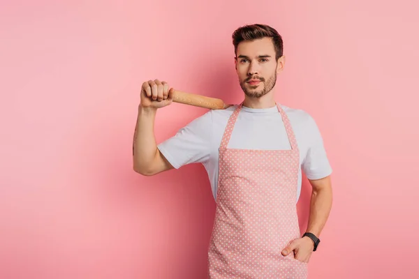 Joyeux jeune homme dans tablier tenant rouleau à pâtisserie tout en se tenant la main dans la poche sur fond rose — Photo de stock