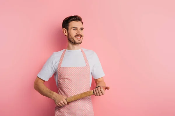 Cheerful young man in apron looking away while holding rolling pin on pink background — Stock Photo